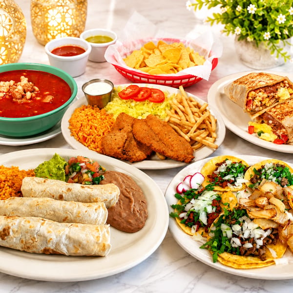 A table laden with various Mexican dishes including tacos, burritos, soup, and fried fish.