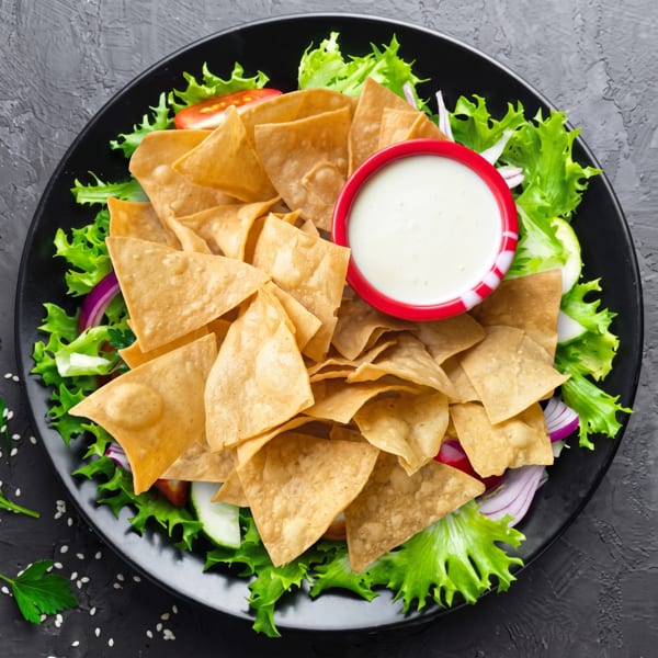 Nachos with tortilla chips, fresh green lettuce, red onion, tomato, cucumber, and a side of white sauce.